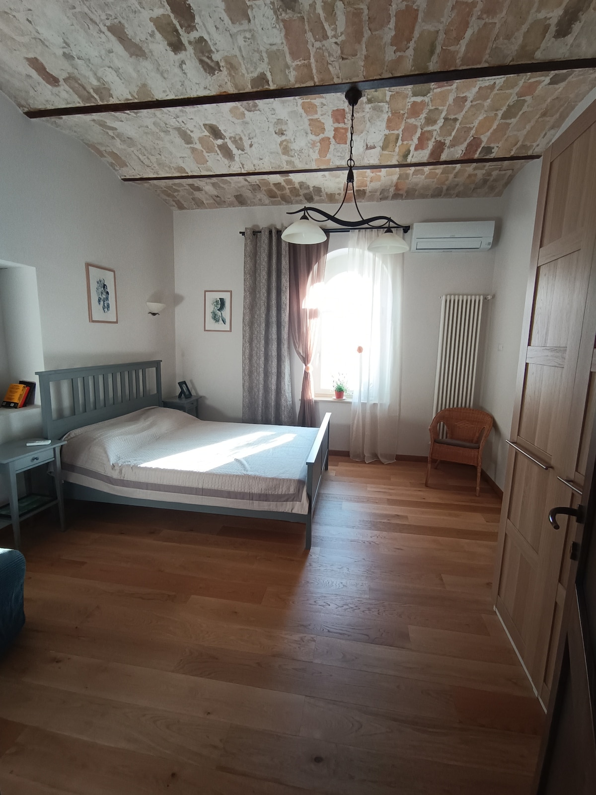 A spacious bedroom features a wooden bed frame with a simple mattress. Light-colored walls are complemented by a rustic ceiling with exposed bricks. A small desk is positioned beside the bed, and a wicker chair sits in the corner near a window dressed with sheer curtains and a light fixture overhead.