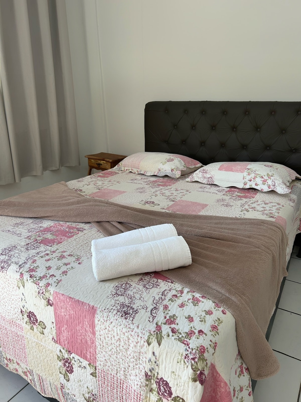 A cozy sleeping area features a queen bed adorned with a patchwork quilt in soft floral patterns. Two pillows rest against a tufted headboard. A light brown towel is neatly placed on a folded blanket at the foot of the bed, and light enters through a nearby window with light curtains.