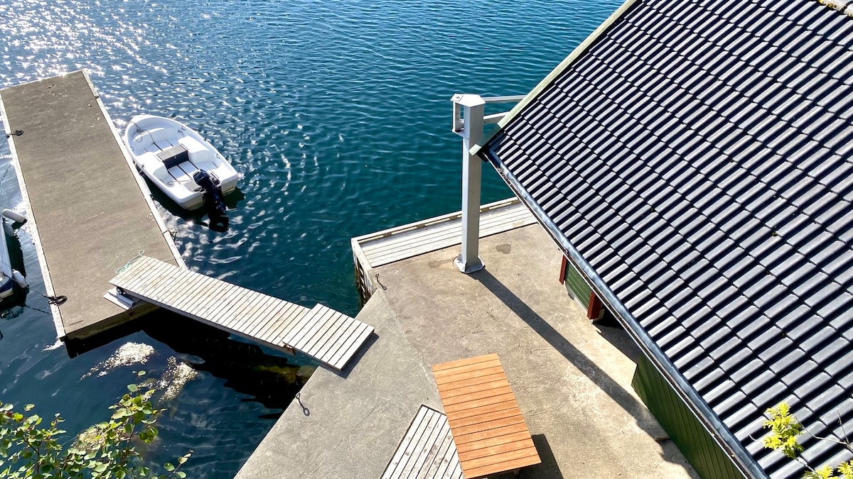 A serene waterside view features a boat dock and a small motorboat tied alongside. A sunlit terrace with a wooden table and chairs overlooks the calm, rippling water. The roof of a nearby structure is visible, reflecting the natural surroundings.