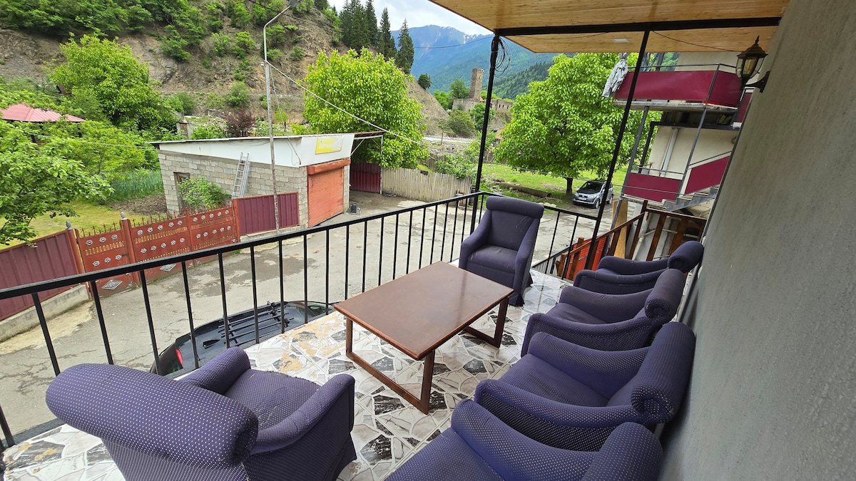 A covered balcony features a wooden table surrounded by six comfortable chairs, offering mountain views and greenery. The railing enhances safety while providing an unobstructed sightline to the road below, where trees and parked vehicles are visible.