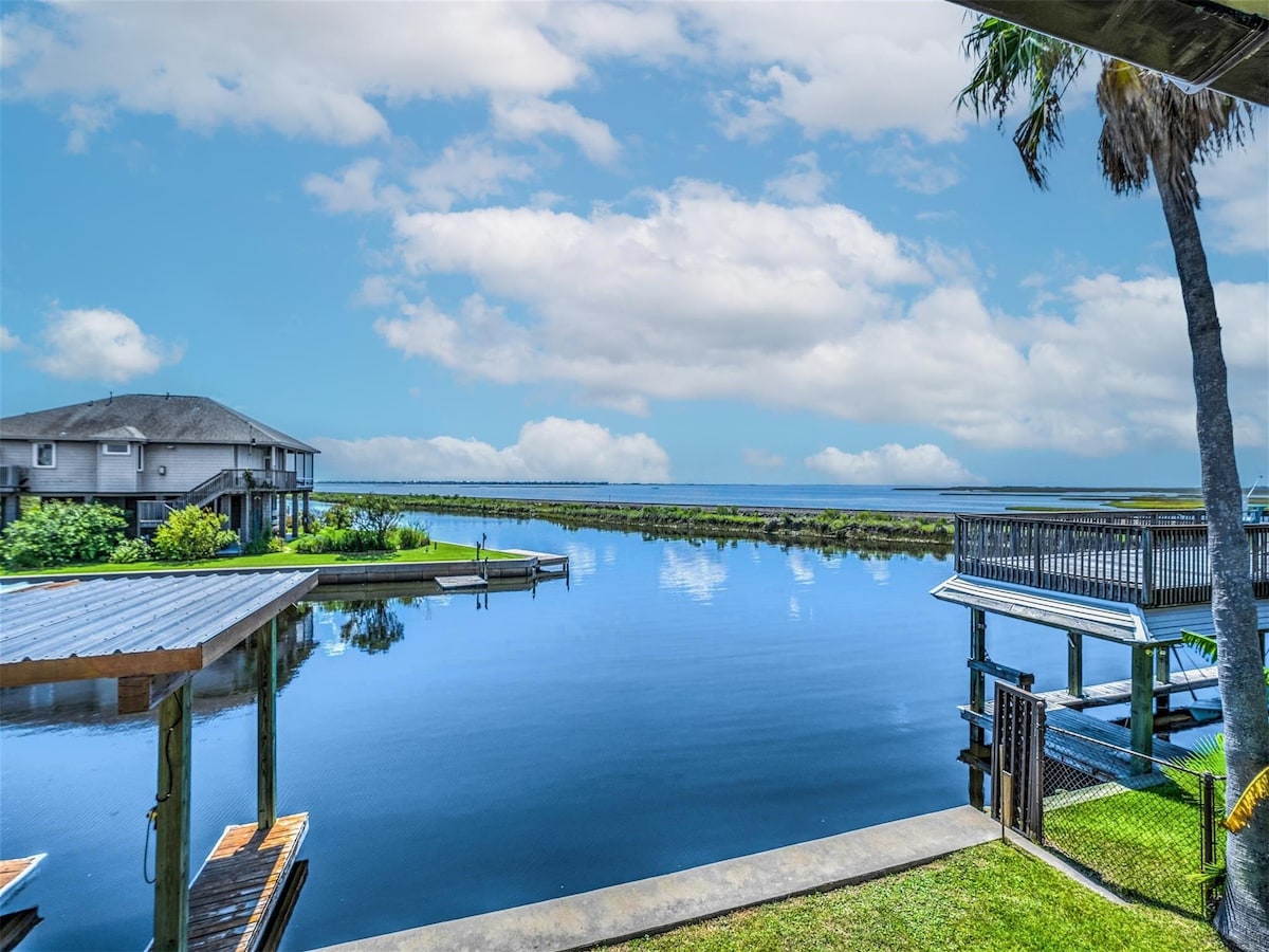 A wide view of a calm waterway is presented, reflecting fluffy clouds and blue skies. Lush greenery borders the banks, enhancing the tranquil scenery. A deck is visible in the foreground, overlooking the water, while two additional structures can be seen in the background.