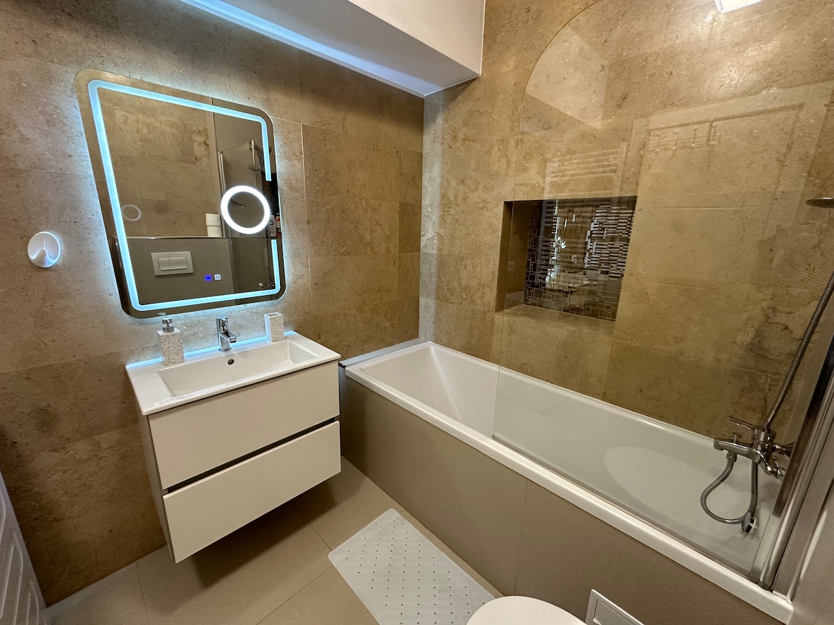 The bathroom features a modern, neutral design with stone-tiled walls and a well-lit mirror. A spacious bathtub is paired with a glass shower enclosure. A sleek vanity with storage space is positioned beside the bathtub, and a white toilet is neatly arranged in the corner.