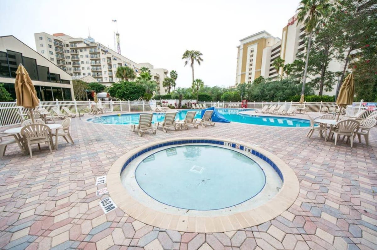 The image features two outdoor swimming pools surrounded by lounge chairs, with an adjacent whirlpool. Trees and tropical plants are visible in the background, enhancing the serene environment. Umbrellas provide shade over the seating area, creating a comfortable space for relaxation.