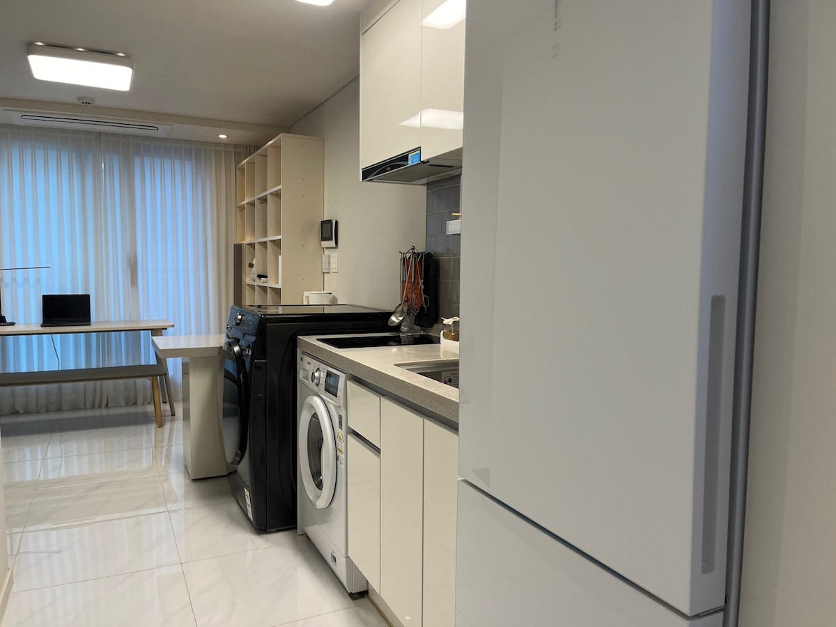 The kitchen area features modern appliances including a refrigerator and a washing machine. A well-organized countertop with cooking utensils is visible. Light filters through the sheer curtains, providing a bright atmosphere. A dining table for three is positioned in the corner.