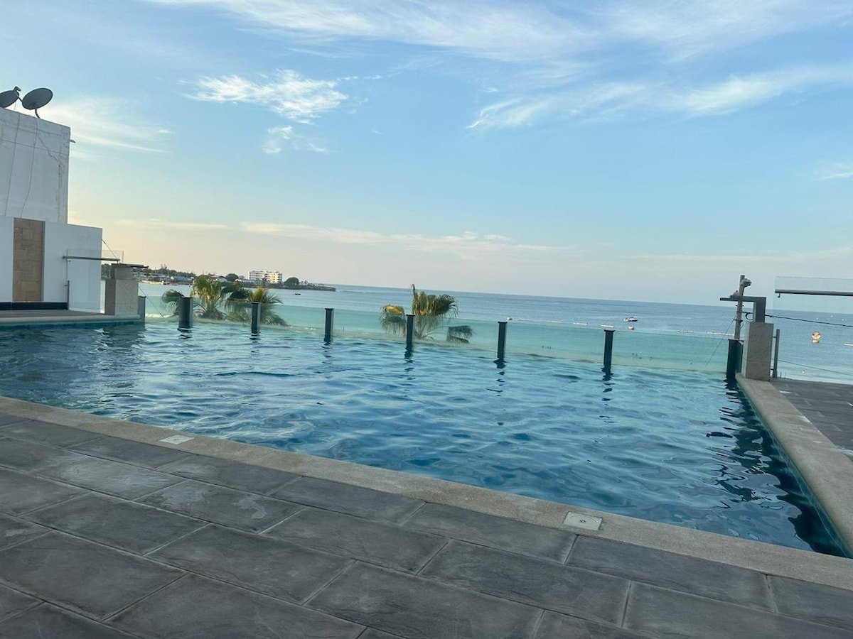 An infinity pool extends towards the ocean, offering unobstructed views of the water. The calm surface reflects the clear sky, with a few palm trees lining the water's edge. The surrounding deck features smooth stone tiles, enhancing the relaxing ambiance.