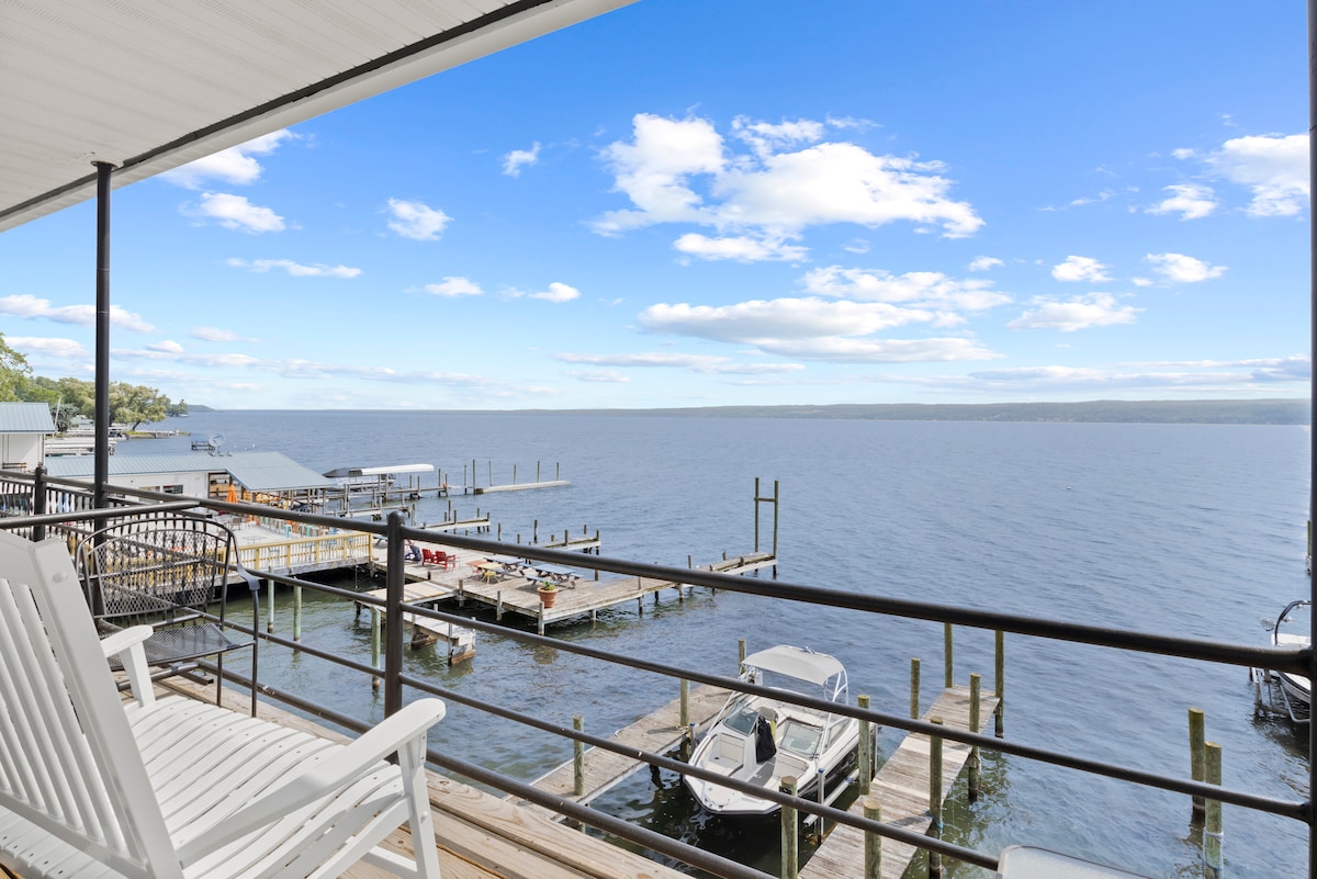 A spacious balcony offers a view of Seneca Lake, with docks and boats lining the water's edge. The sky is bright and clear, reflecting on the lake's surface. Comfortable seating is provided for enjoying the serene surroundings.