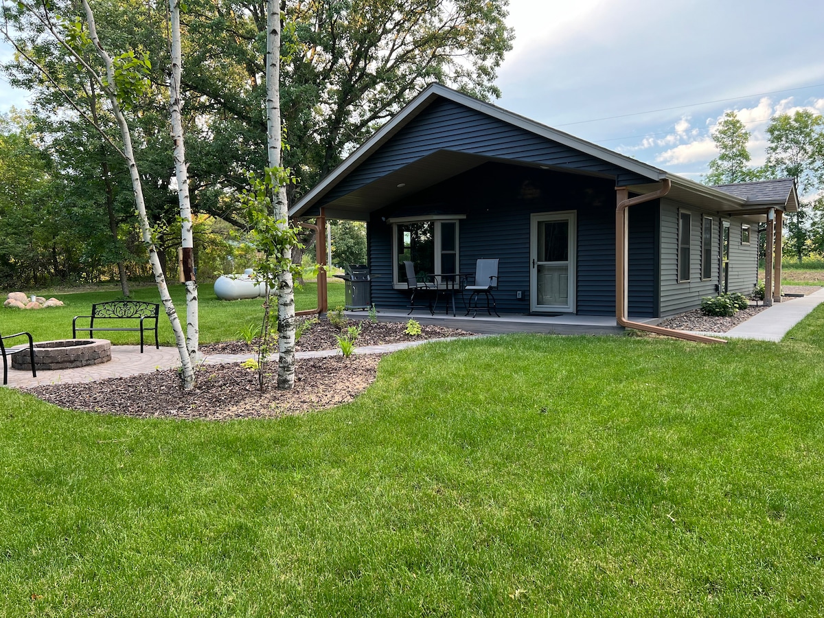 A newly built cottage features a dark blue exterior and a covered porch with seating. Gravel pathways lead through green grass, flanked by landscaped areas with greenery and stone accents. Trees provide shade, creating a serene outdoor space for relaxation.