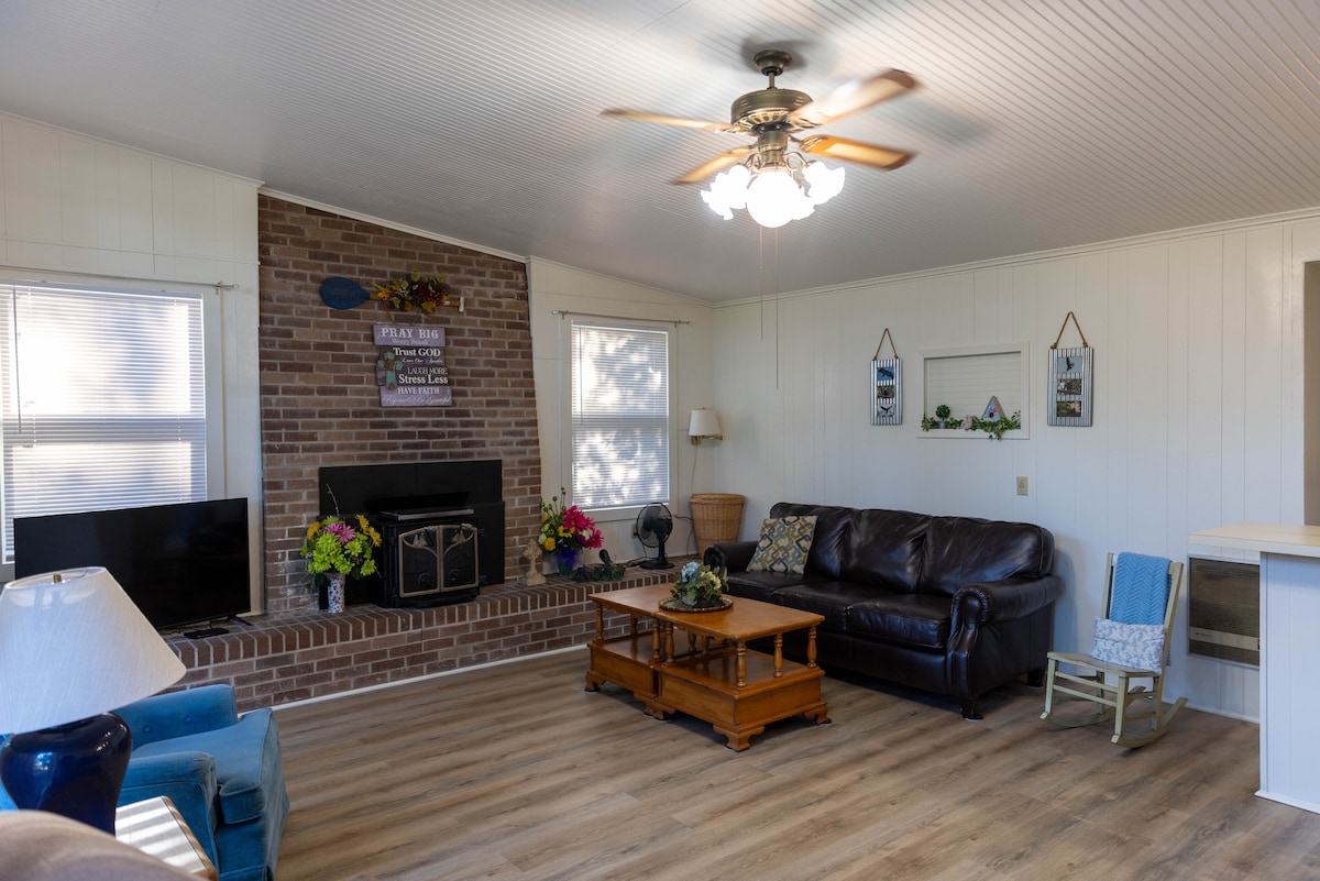 A spacious living area features wood flooring and a ceiling fan. A brick fireplace is highlighted by decorative items and fresh flowers. Comfortable seating includes a dark leather couch and a wooden coffee table. Large windows allow natural light to fill the space.