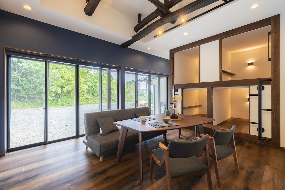 A bright living space features large glass doors that invite natural light and views of greenery. A comfortable gray sofa and a wooden dining table are central to the layout. A cozy nook with sliding doors is visible, offering a playful, intimate atmosphere.