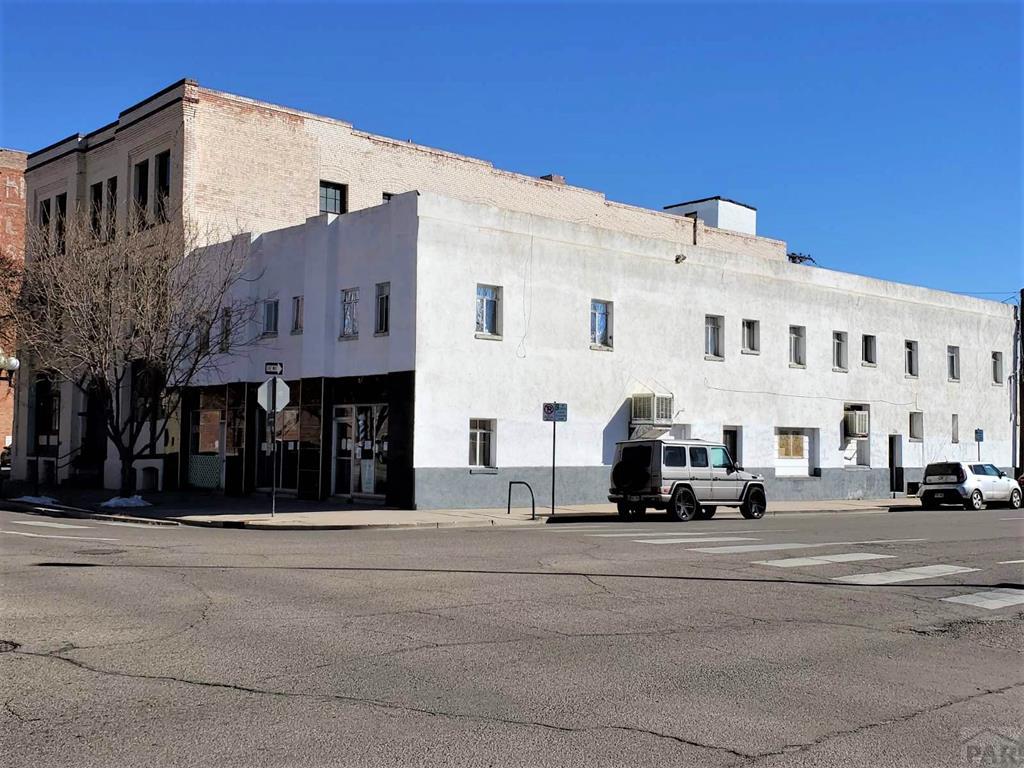 A three-story building with a white exterior and a simple architectural design is depicted. The ground floor includes large windows and entry points. Two vehicles are parked nearby, and street signs can be seen at the intersection, highlighting the urban setting.