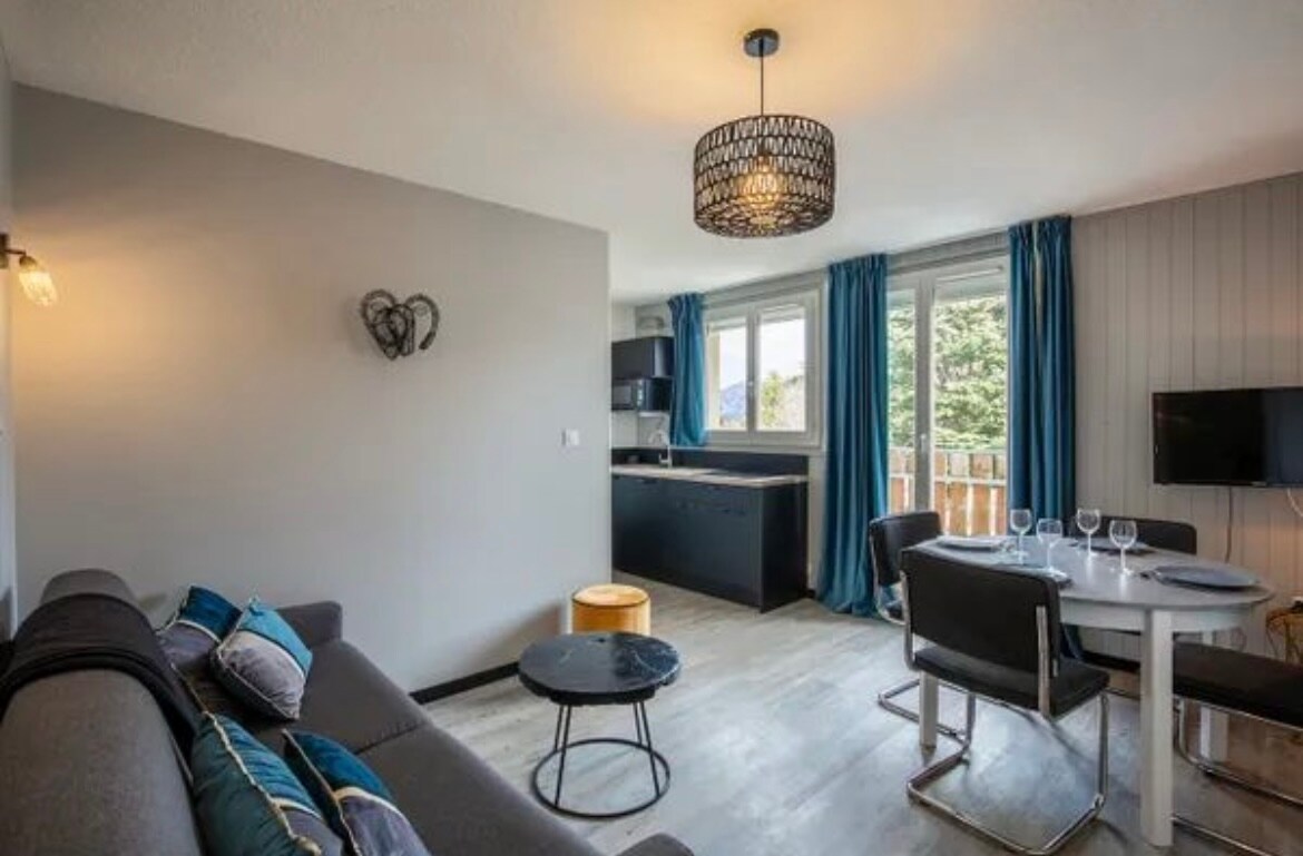 An open living space is seen, featuring a comfortable sofa adorned with decorative pillows. A round dining table set for four is positioned beside a window, where blue curtains complement the contemporary decor. A kitchenette is visible, with modern appliances included.