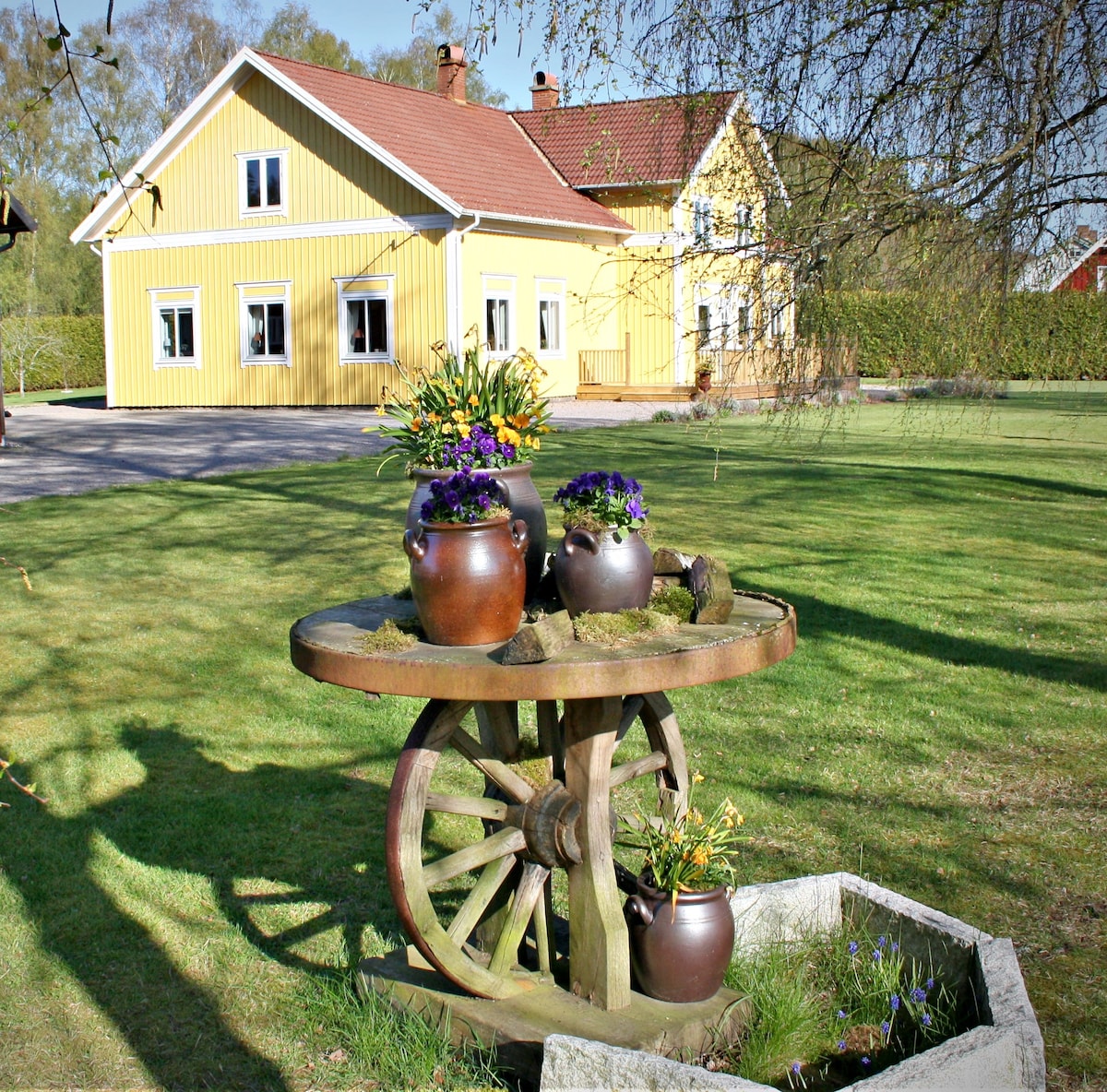A rustic wooden wheel table is featured in the foreground, adorned with brown pots filled with blooming flowers. The vibrant yellow exterior of the house, complemented by multiple windows, is visible in the background, surrounded by a well-maintained lawn.