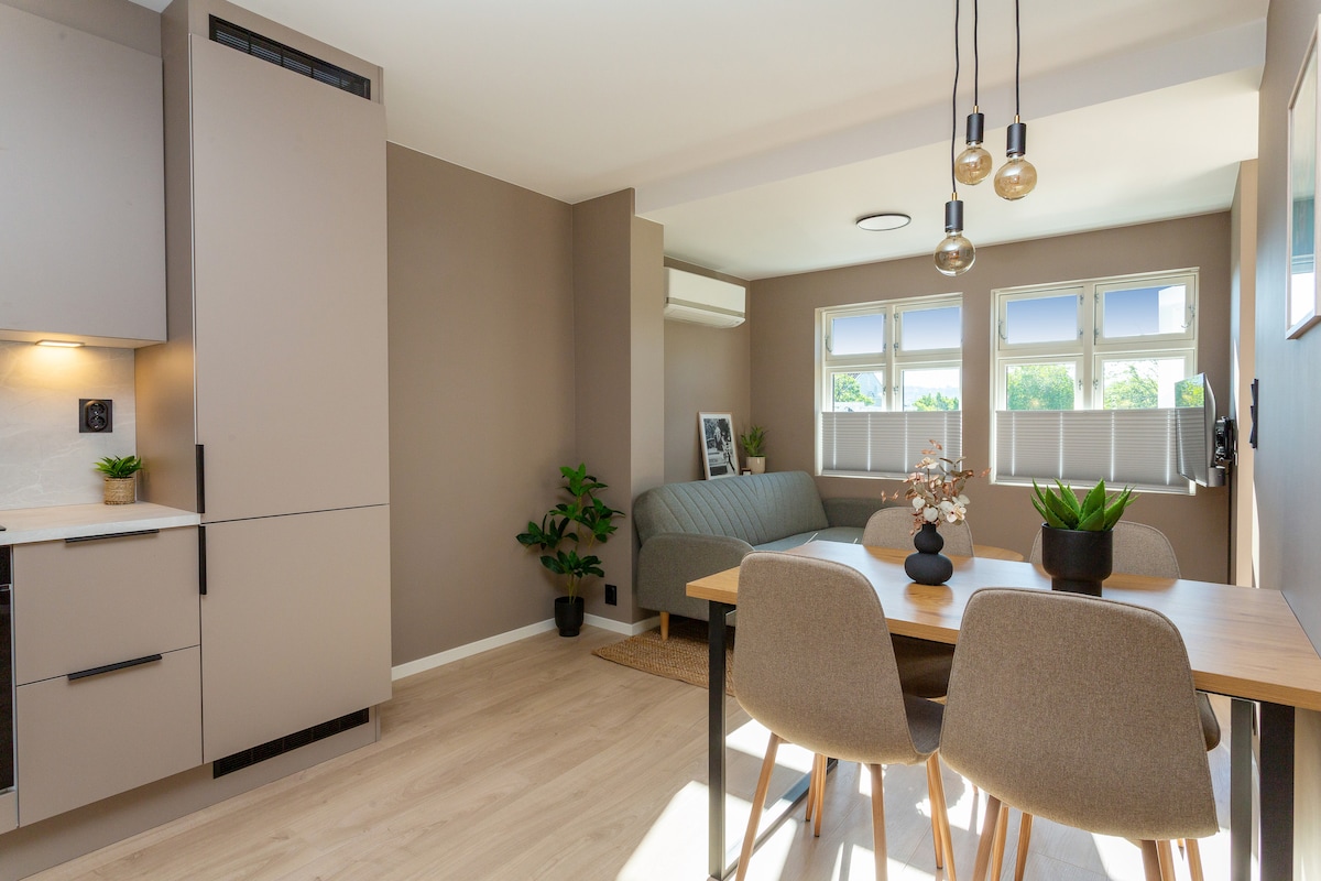 The open living area features a modern kitchen with a light-colored cabinetry and a spacious dining table accommodating four chairs. Soft natural light fills the room through multiple windows, complemented by indoor plants for a refreshing touch.
