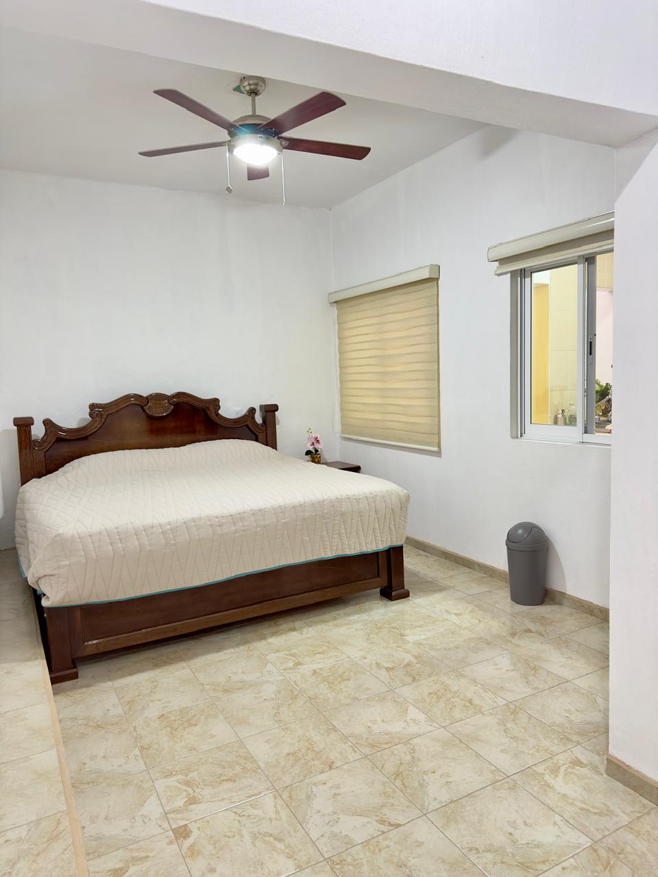 A spacious bedroom features a large wooden bed with a light-colored quilt. A ceiling fan is mounted above, and natural light enters through the window adorned with blinds. A wastebasket is located near the corner, complementing the neutral tone of the tiled floor.