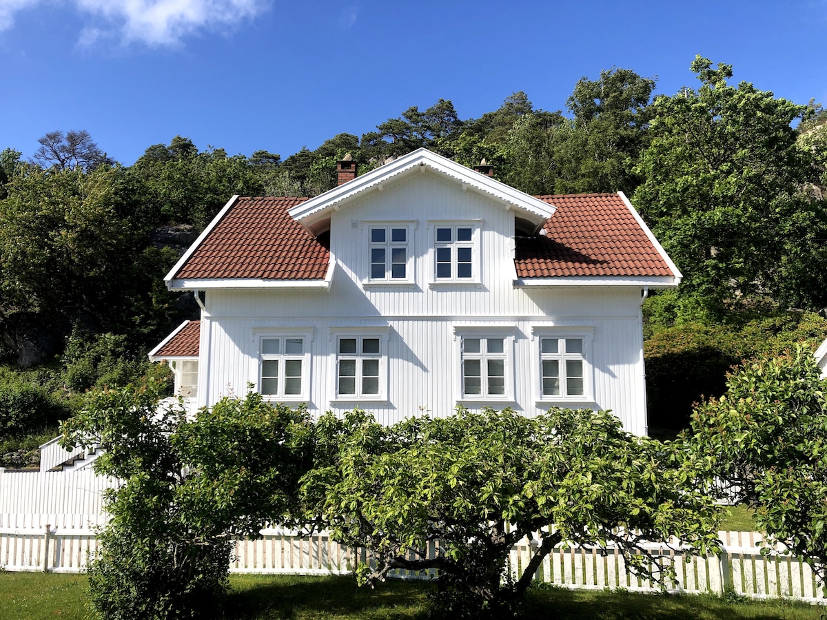 The charming white skipper house, framed by greenery, features a red roof and multiple windows that invite natural light. A white picket fence encircles the property, enhancing the feeling of a peaceful retreat in a natural setting.