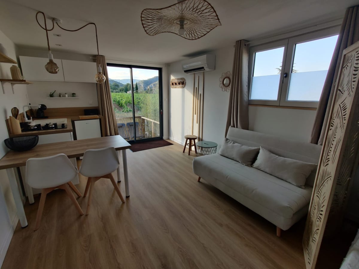 A light-filled living space features a small dining table with two chairs and a cozy sofa. The kitchen area is designed with modern cabinetry and includes essential appliances. Large windows open to a view of greenery, adding a sense of connection to nature.