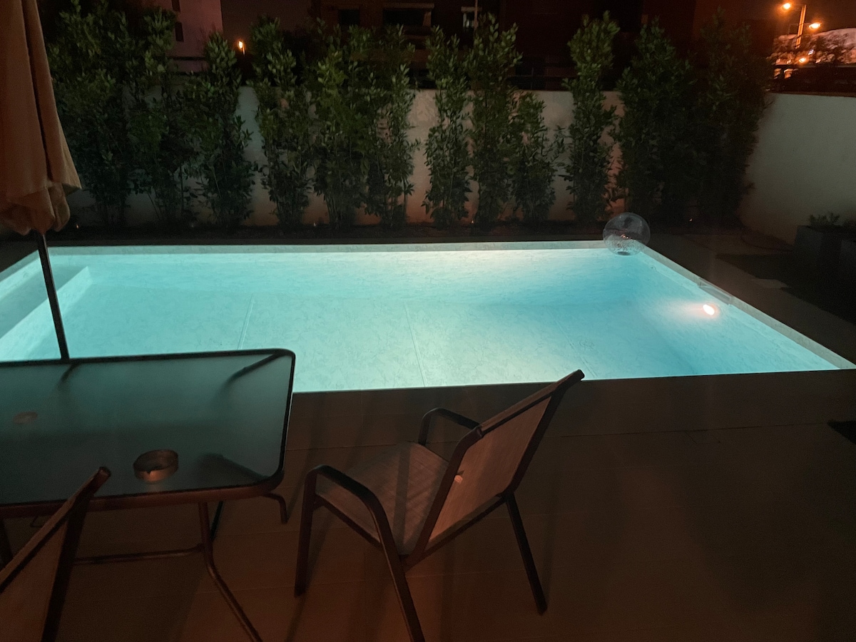 A serene pool area is presented, featuring a softly illuminated water surface. Surrounding greenery adds privacy, while a simple table and chair are positioned nearby for relaxation. The tranquil ambiance is highlighted by the gentle glow of the pool light against the evening backdrop.