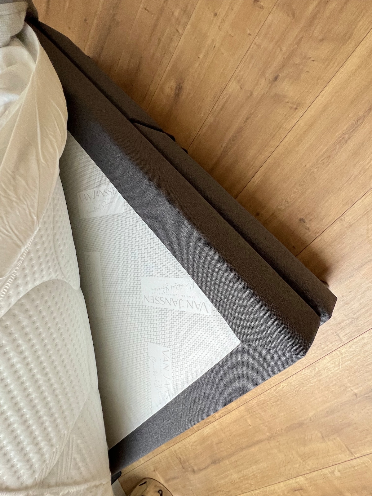 A spacious mattress is depicted, resting on a modern bed frame with a soft gray fabric finish. The natural wood floor complements the warm tones of the room, while a textured white mattress protector is visible, showcasing its quilted design.