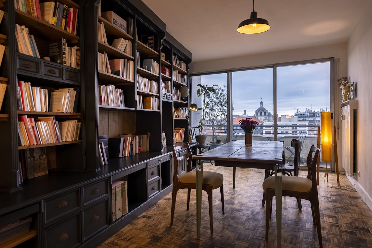 A spacious dining area features a wooden table surrounded by chairs. A large bookshelf filled with books occupies one wall, while a large window offers views of the cityscape. A plant and a flower pot add a touch of greenery, complemented by soft lighting from a pendant lamp.