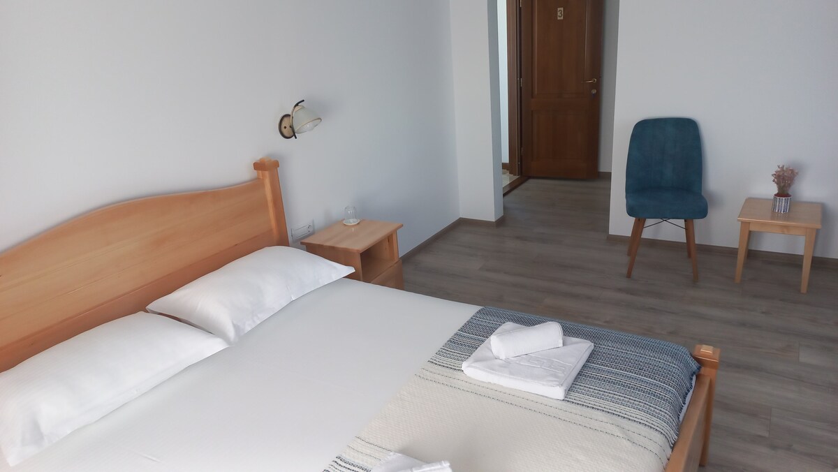 A minimalist bedroom features a spacious bed dressed with white linens and a decorative throw. A light wood bedside table holds a glass of water. A blue chair and small wooden table are positioned nearby, with a door leading to another area visible in the background.