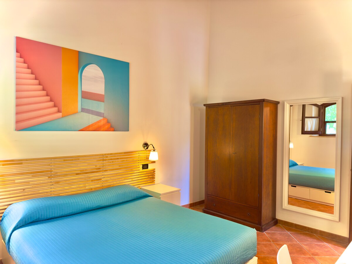 A spacious bedroom features a large bed dressed in a textured blue cover, complemented by a modern wooden headboard. A colorful artwork of staircase and arch decorates the wall above. A large mirror and a dark wooden wardrobe are also included, enhancing the room's functionality.