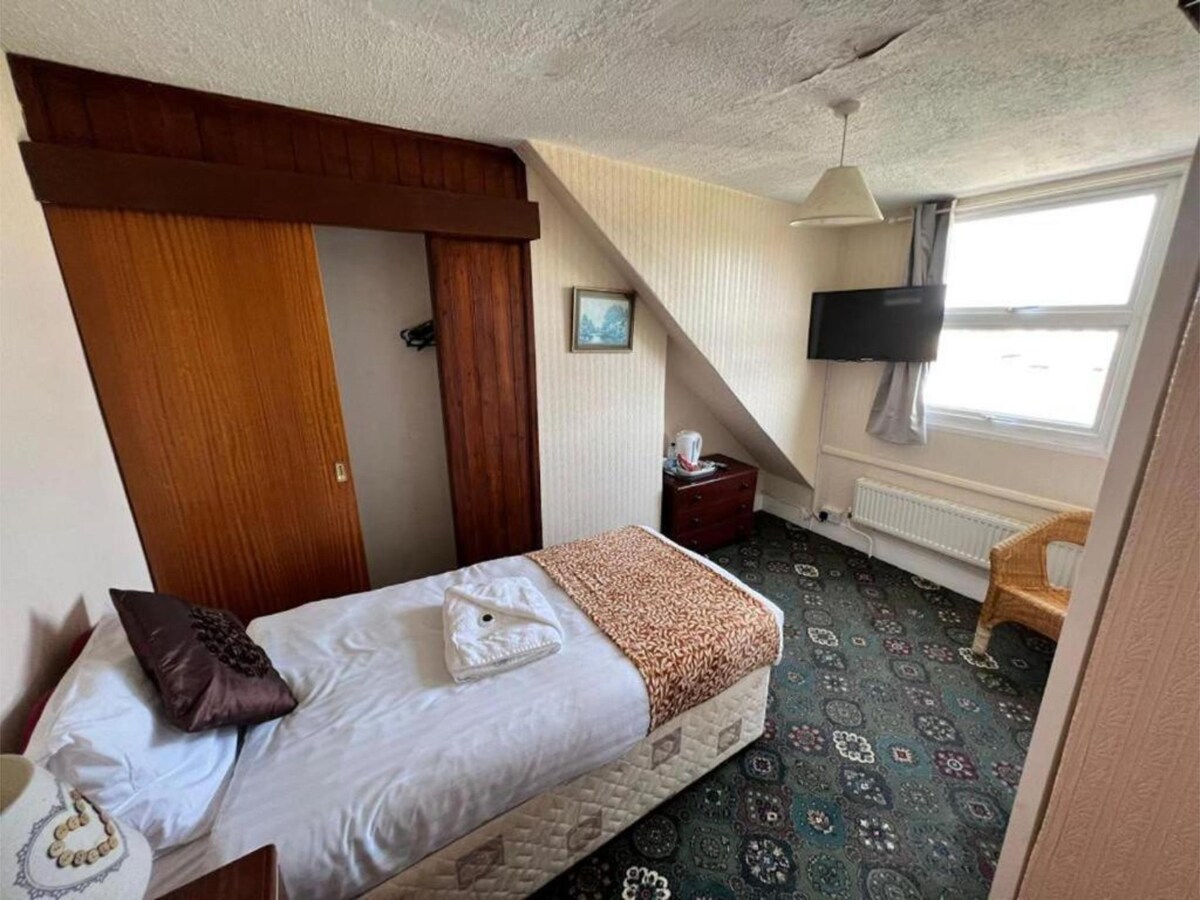 A cozy room features a single bed with a warm-toned throw and neatly arranged towels. A wooden wardrobe and a dark wood dresser provide storage. Natural light enters through a window adorned with light curtains, and a wall-mounted TV is positioned for viewing.