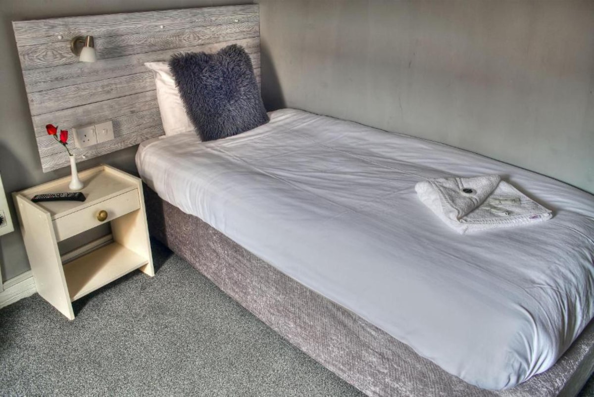 A single bed is set against a textured headboard, featuring neutral bedding arranged neatly. A bedside table holds a small vase with a rose and a soft towel is placed at the foot of the bed. Soft grey carpeting underlines the relaxing atmosphere.