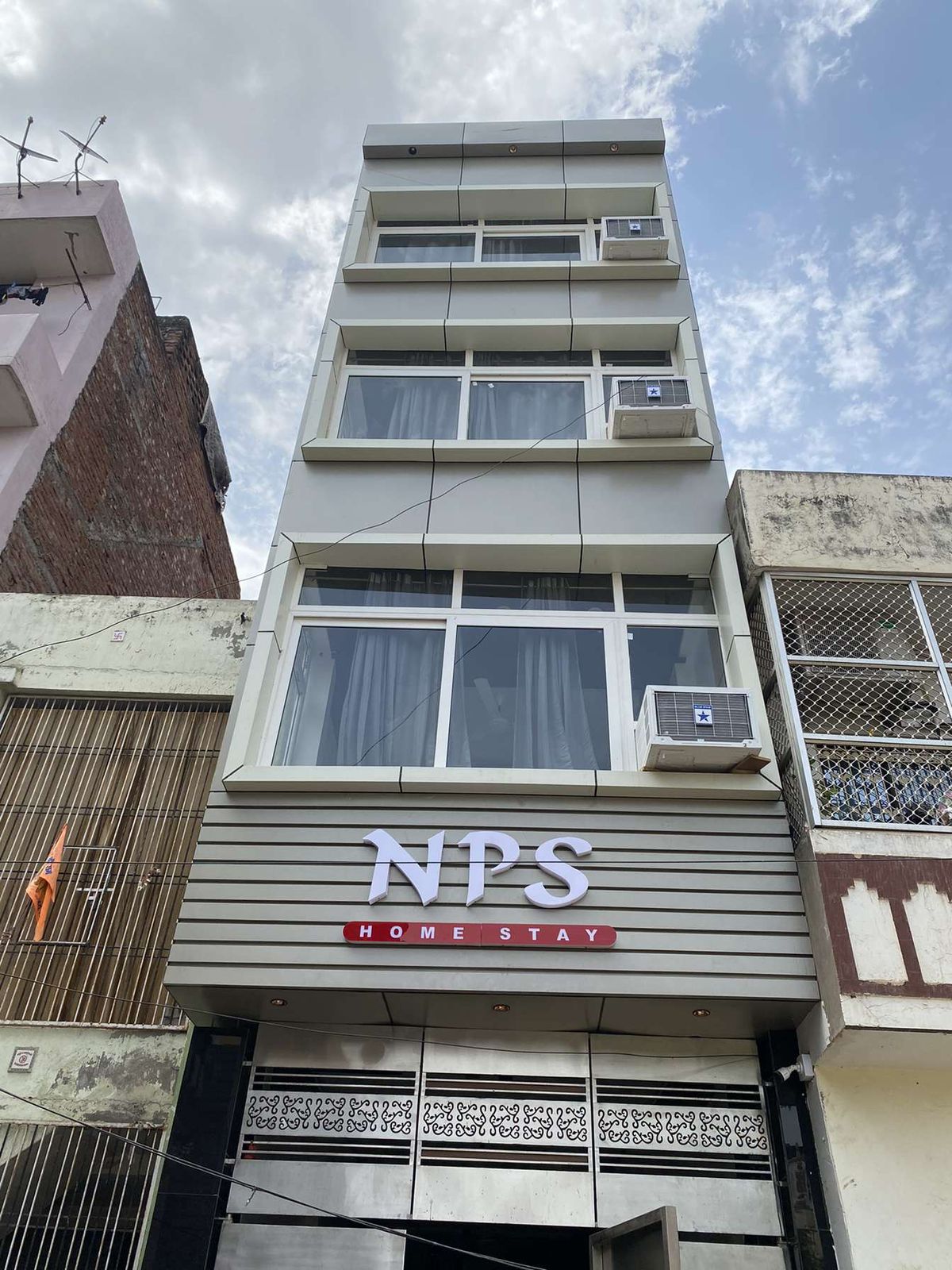 The exterior of NPS Home Stay is captured, showcasing a modern four-story building with large glass windows. The name 'NPS' is prominently displayed in white against a gray facade. Air conditioning units are installed on several levels, blending functionality with a contemporary design.