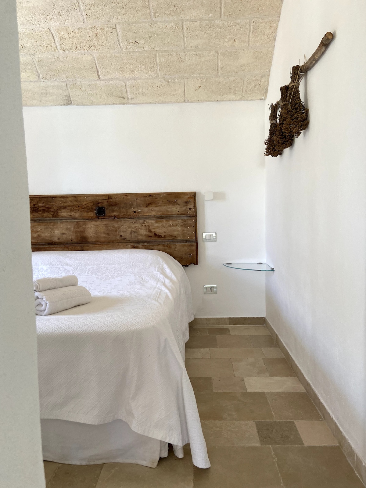 The suite features a welcoming sleeping area with a spacious bed adorned with white linens. Natural stone walls and a light-colored tile floor complement the rustic wooden headboard. Soft towels are neatly arranged, creating an inviting ambiance.