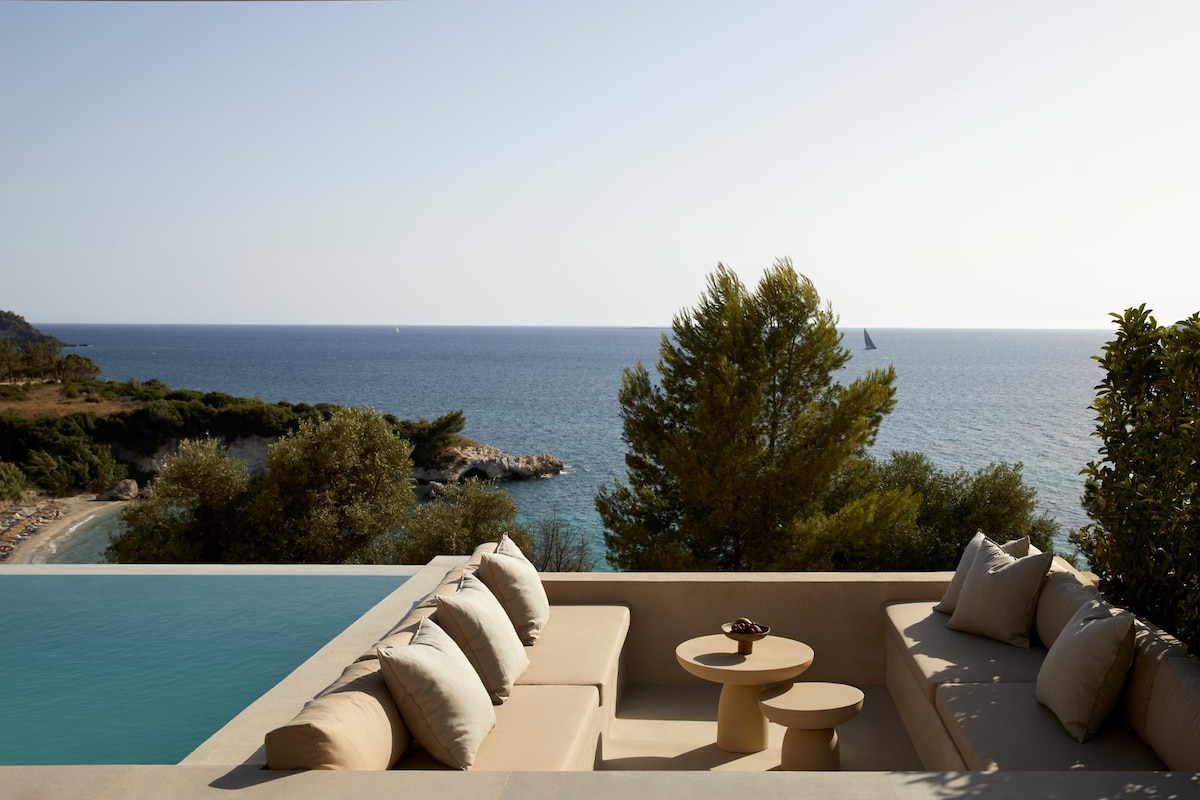 An outdoor seating area features a low sofa adorned with cushions, paired with a round table. The space overlooks a serene expanse of the Ionian Sea, enhancing the coastal ambiance. A backdrop of greenery adds to the natural setting surrounding the pool.