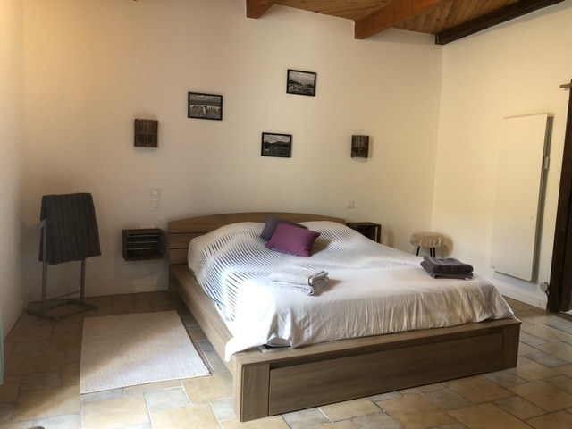 A spacious bedroom features a large bed with a white cover and a decorative pillow. The room's stone floor is complemented by a soft area rug. Wall-mounted decorations and a standing mirror add character, while natural light illuminates the neutral-toned walls.