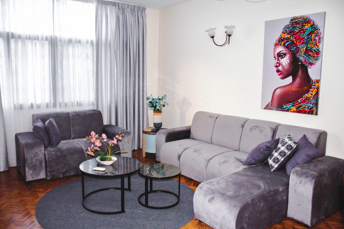 A modern living room is shown, featuring a plush gray sofa and matching chairs arranged around two coffee tables. A round area rug anchors the seating. Natural light filters through sheer curtains, illuminating a colorful artwork on the wall and potted plants accentuating the space.