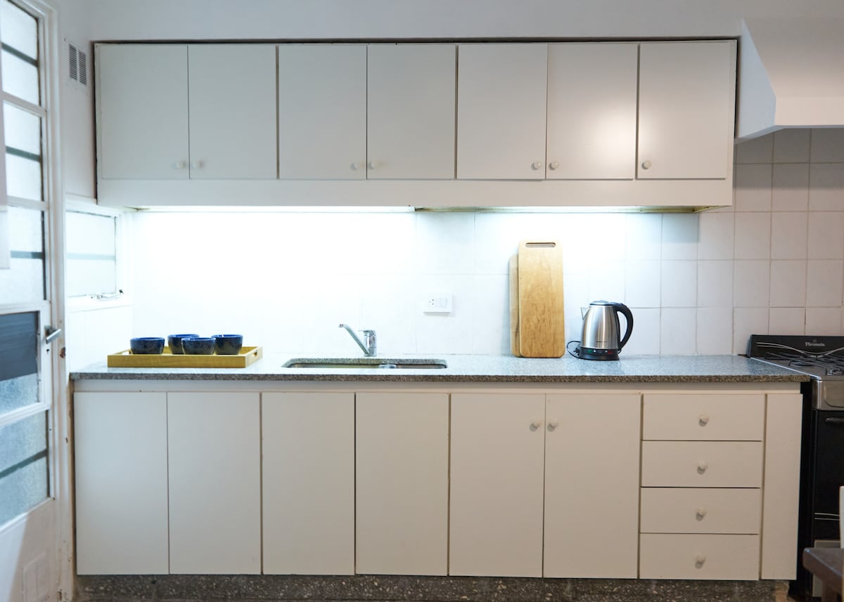 The kitchen features sleek, white cabinetry with overhead storage and a spacious countertop. A kettle is positioned next to a wooden cutting board, with dishes placed on a tray. Soft lighting highlights the area, creating a bright and functional workspace.