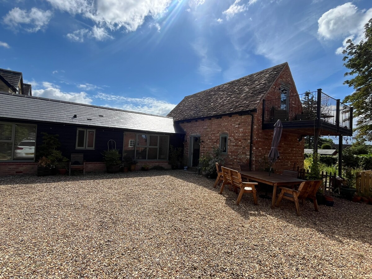 The exterior of the Old Barn reveals a blend of brick and wood finishes beneath a clear blue sky. A spacious gravel area is visible, with a large wooden dining table and chairs positioned for outdoor dining. A separate balcony area can be seen above.