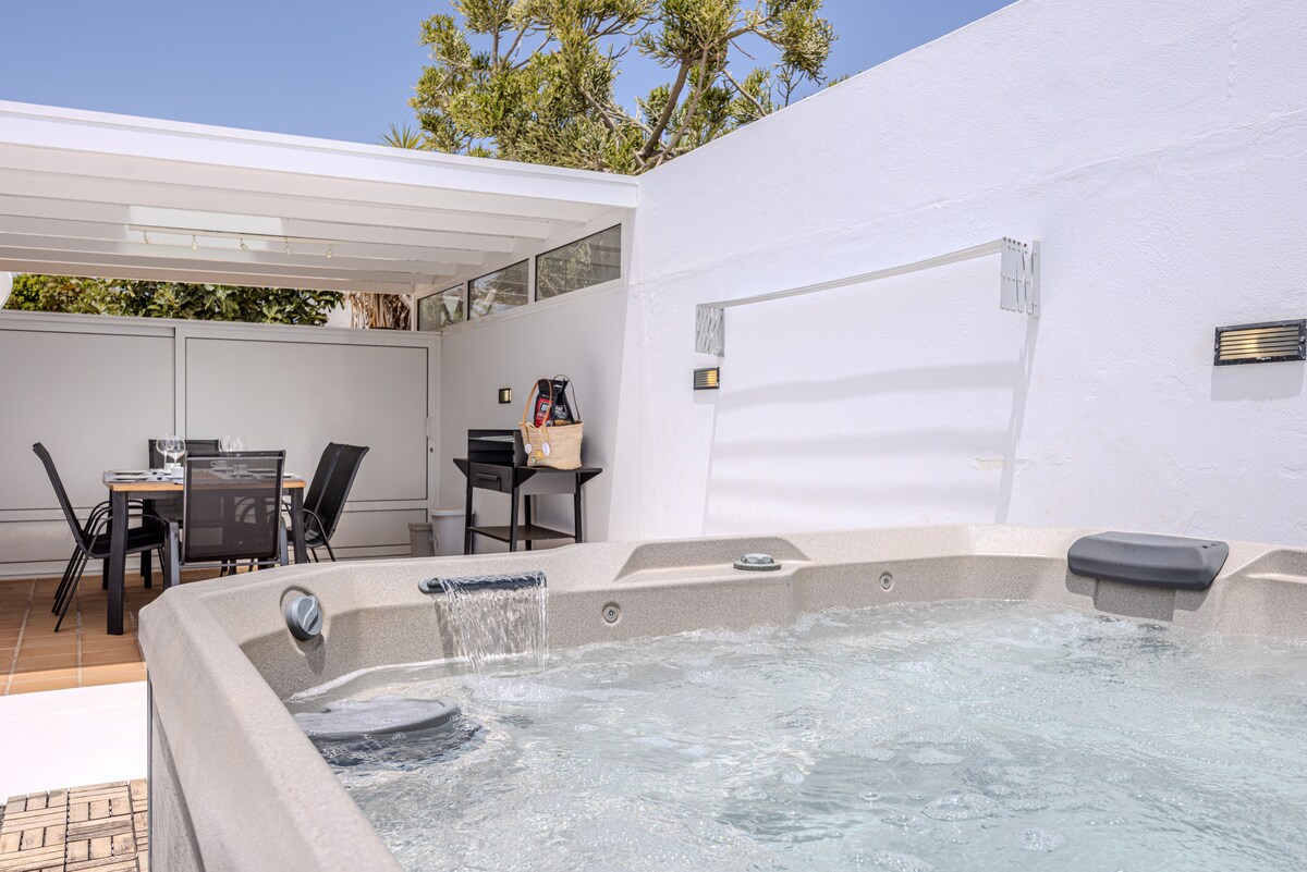 An outdoor jacuzzi is featured in a sunlit patio, with water gently bubbling. In the background, a dining area is visible, equipped with a table and chairs under a sheltered space, surrounded by greenery.
