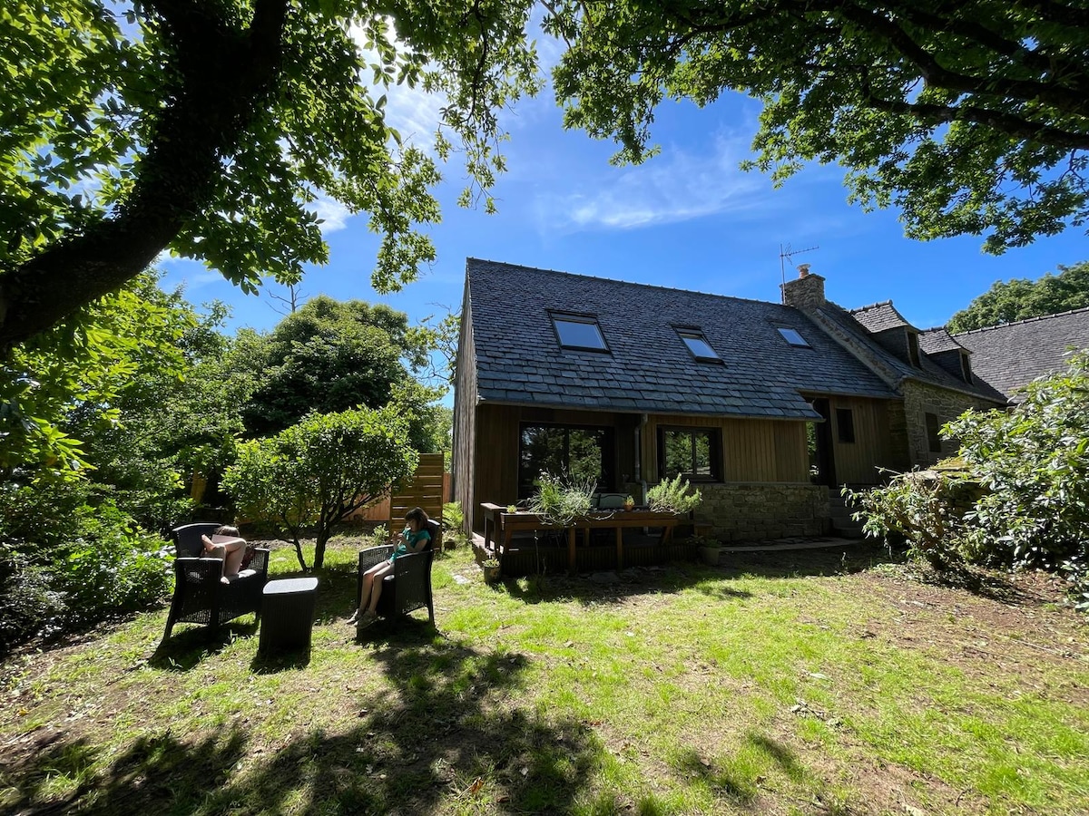 A charming house is set amidst lush greenery, featuring a sloped slate roof and large windows. A small outdoor seating area with two chairs is visible on the lawn, surrounded by low shrubs and trees, providing a serene environment.