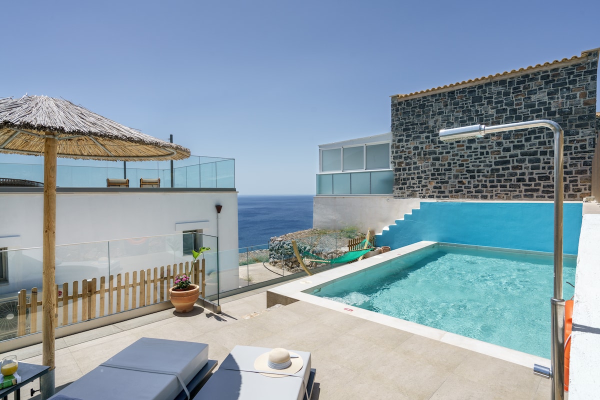 An outdoor area features a refreshing swimming pool next to a sun terrace, equipped with comfortable loungers and a thatched umbrella for shade. The backdrop includes a stone wall and views of the serene sea, creating a peaceful atmosphere for relaxation.
