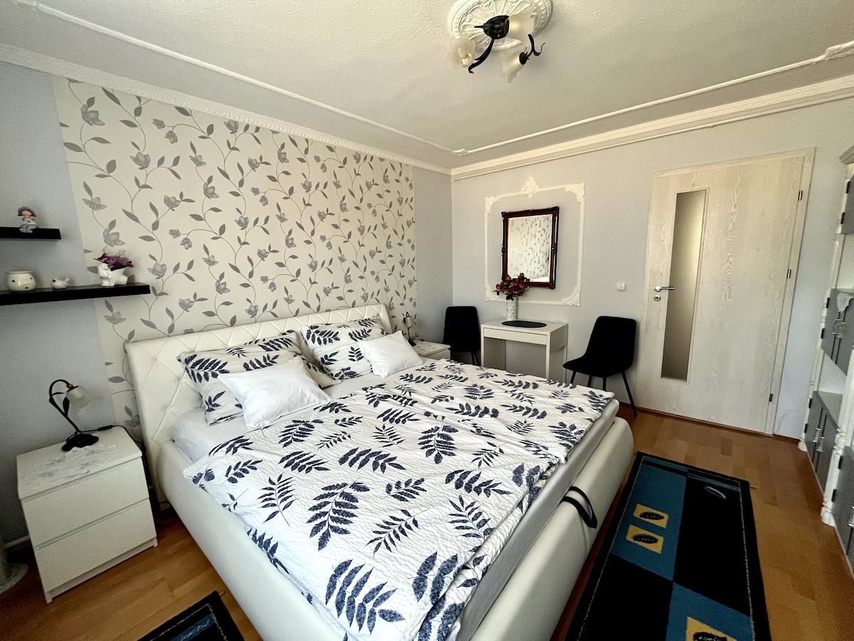 A spacious bedroom features a large bed with a floral-patterned blanket, complemented by pillows. A dark wood mirror hangs above a minimalist desk, while two chairs are positioned nearby. Light-colored walls are adorned with a leafy wallpaper design, creating a serene environment.