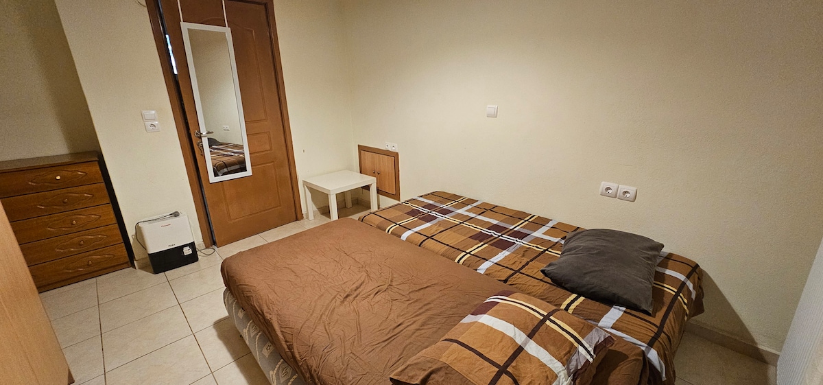 A simple sleeping area is presented, featuring a spacious bed with a brown checkered blanket. A neutral pillow rests on the bed. A small white table and a wooden dresser with multiple drawers are also visible, along with a door showing a mirrored panel.