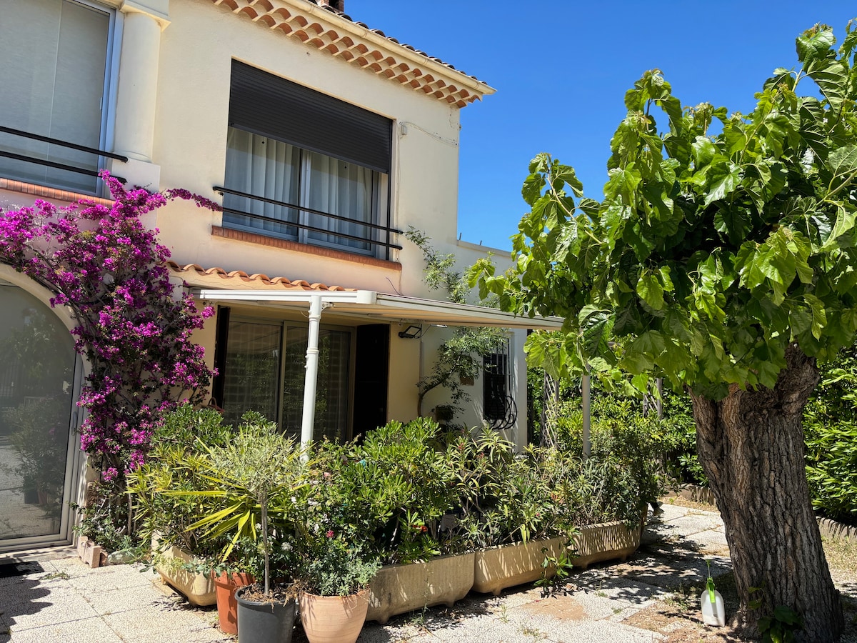 A serene villa exterior is surrounded by lush greenery and colorful flowering plants. A shaded terrace provides a relaxing spot, while large windows allow natural light to flood the interior. A mature tree adds character to the inviting outdoor space.