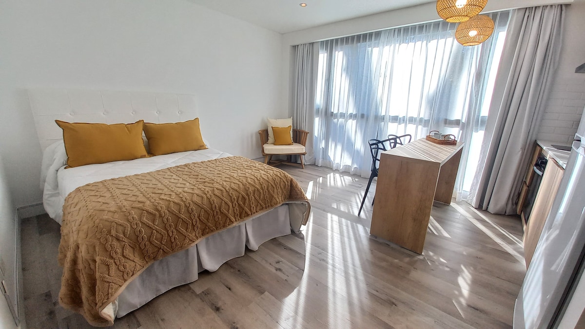 A bright and spacious studio features a queen-sized bed with a textured blanket and two decorative pillows. A cozy armchair is positioned next to a wooden table, creating a functional area. Large windows adorned with sheer curtains allow natural light to enhance the room's warmth.