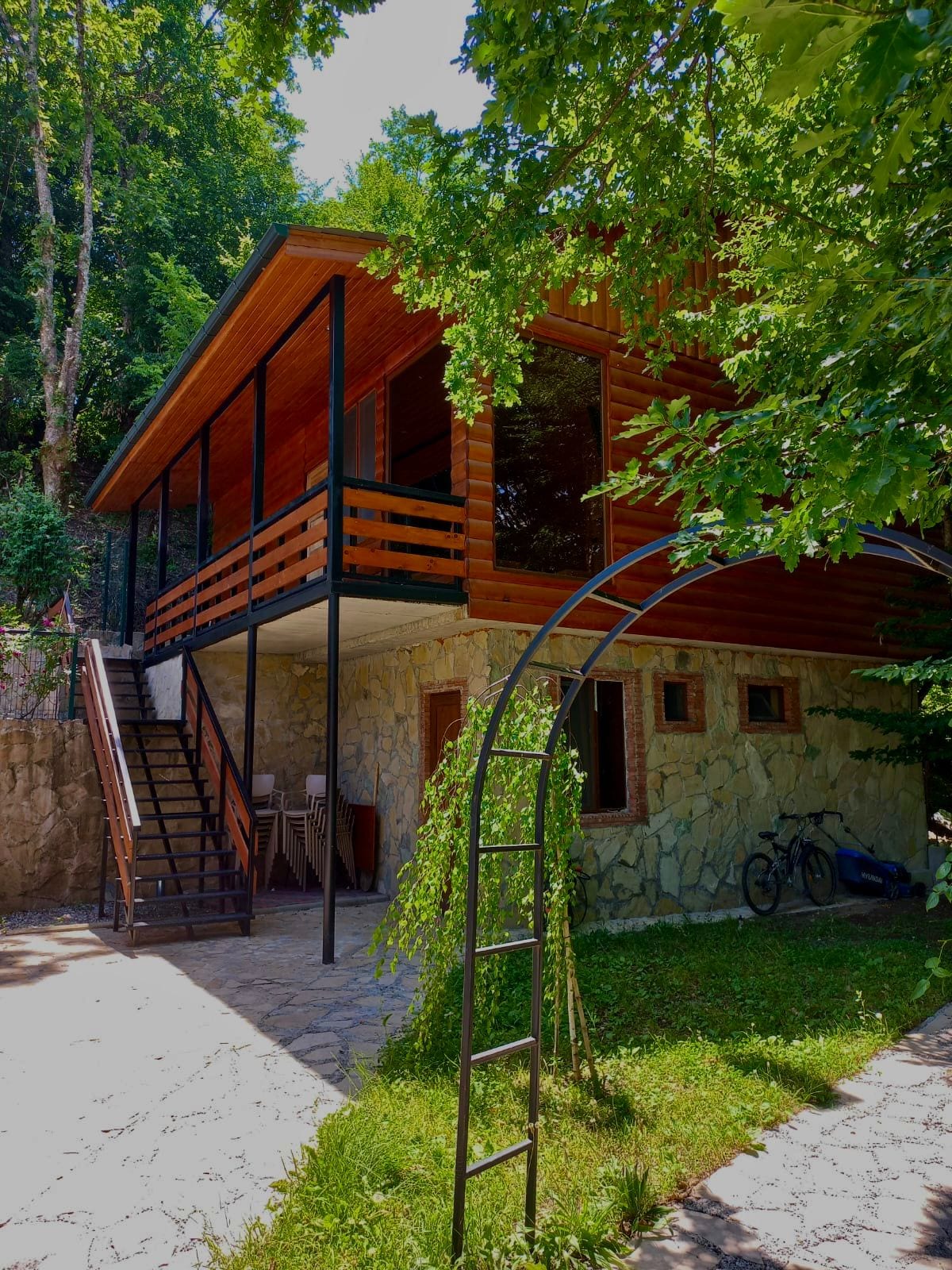 The cottage is surrounded by lush greenery, featuring a wooden upper deck with a railing. A staircase leads to the entrance, while a stone base is visible beneath. A bicycle is parked near the building, enhancing the natural, outdoor setting.
