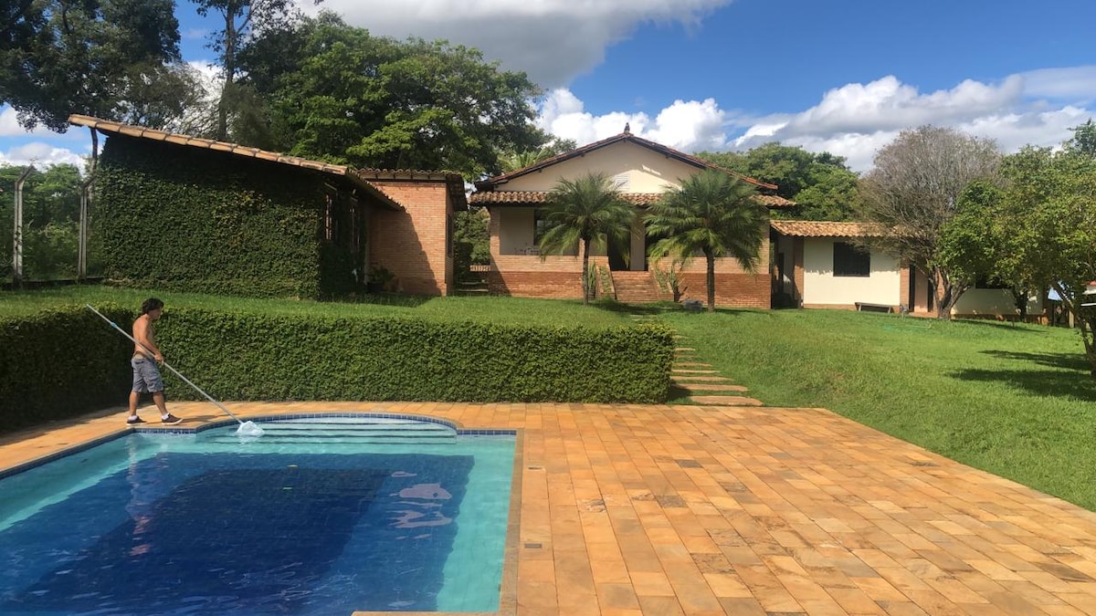 A serene outdoor space features a clear swimming pool, with a stone patio extending around it. Lush green grass and neatly trimmed hedges surround the pool area, while a charming house with a tiled roof and palm trees is visible in the background under a blue sky.