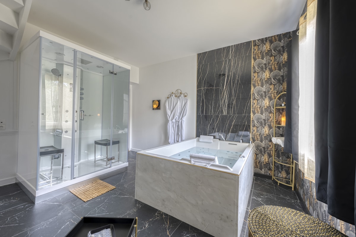 The spa area features a luxurious whirlpool tub and an oversized shower cabin with glass doors. Elegant dark marble flooring contrasts with light walls adorned with floral accents. A plush bathrobe hangs nearby, completing the serene ambiance of this relaxation space.