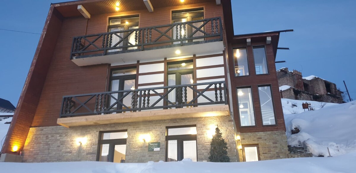A modern three-story building features wooden accents and expansive glass windows. Balconies with decorative railings provide outdoor views. Snow blankets the ground, creating a serene winter scene. Warm lighting illuminates the entrance, highlighting the stone facade and enhancing the building's inviting presence.