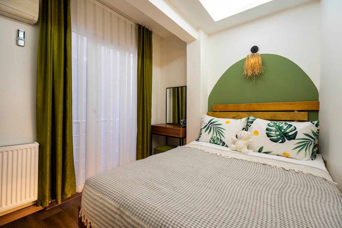 A cozy economy double room features a spacious bed adorned with leaf-patterned pillows and a soft throw. Natural light filters through sheer curtains, highlighting the green accent wall. A wooden desk and mirror are positioned against one wall, adding functionality to the room.
