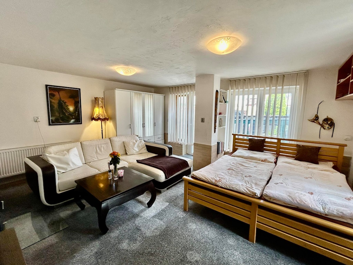 The spacious room features a comfortable seating area with a sectional sofa and a wooden coffee table. A sleeping area with a double bed is present, while a wardrobe offers storage space. Natural light filters through the vertical blinds, creating a bright atmosphere.
