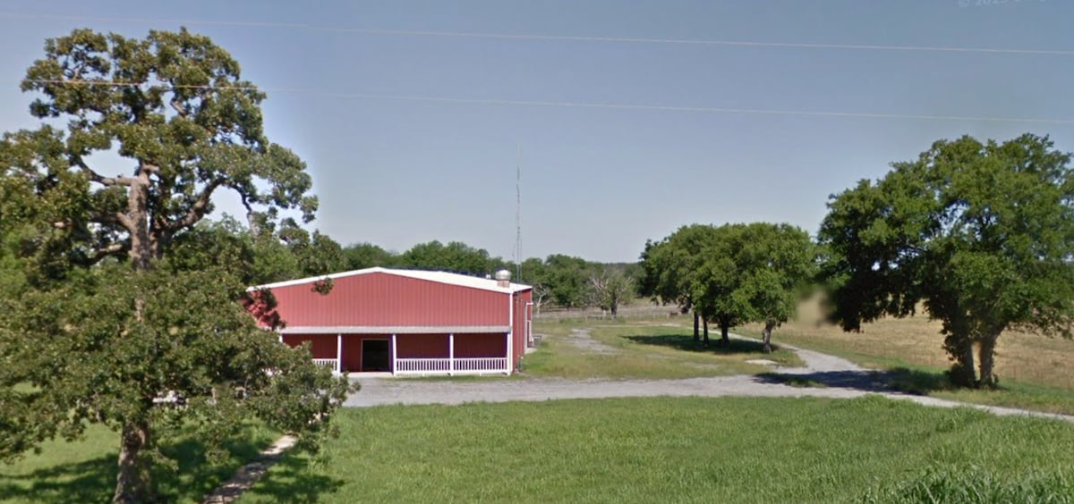 A red barn stands surrounded by grassy areas and trees. The structure features a broad entrance and ample windows that reflect its rustic character. The landscape includes open fields and additional trees, contributing to the serene rural setting.