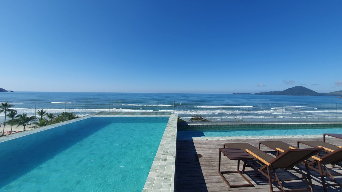An infinity pool extends towards the horizon, offering uninterrupted views of the ocean and distant mountains. Sun loungers are positioned along the wooden deck, providing a space to relax under the clear blue sky, framed by gentle waves and a sandy beach.