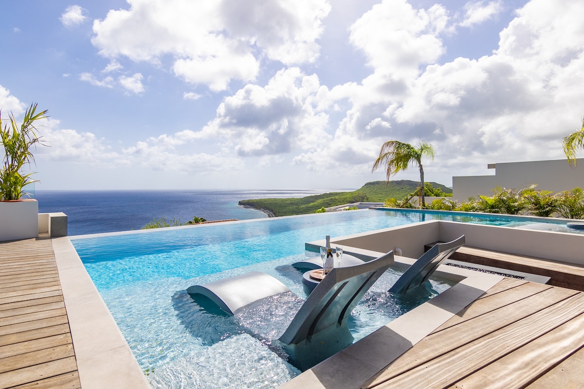 An infinity pool extends towards the horizon, featuring four loungers partially submerged in the water. Lush greenery surrounds the pool area, while the expansive view of the Caribbean Sea and sky creates a serene backdrop.
